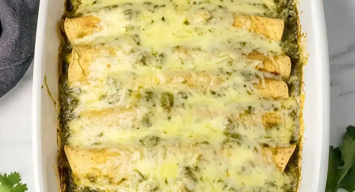 A close-up overhead shot of a white baking dish filled with The Best Green Chili Chicken Enchiladas Recipe, featuring golden-brown tortillas smothered in a creamy green chili sauce and melted white cheese.
