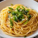 An overhead shot of a light-colored bowl brimming with Vietnamese Garlic Noodles, generously garnished with vibrant green scallions and sesame seeds, resting on a patterned napkin with chopsticks to the right.
