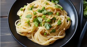 An overhead view of a black bowl filled with Vietnamese Garlic Noodles, brightened with green peas, fresh cilantro sprigs, and chopped peanuts. Dark chopsticks are positioned on the right side of the bowl, set against a dark wooden table.