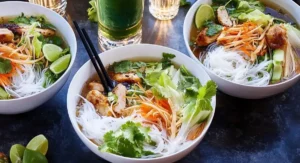Three bowls of Vietnamese Noodle Bowl with Chicken on a dark table with a tall glass of green liquid and lime wedges.
