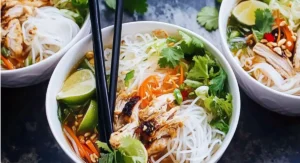 Three white bowls filled with Vietnamese Noodle Bowl with Chicken, featuring rice noodles, grilled chicken, fresh herbs, lime wedges, and broth on a dark surface.