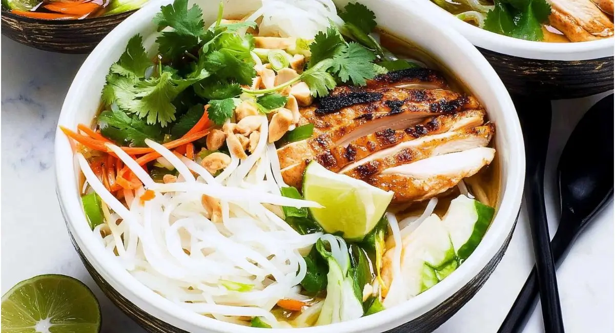 A white bowl of Vietnamese Noodle Bowl with Chicken topped with grilled chicken, herbs, vegetables, a lime wedge, and chopsticks.