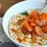 A white bowl of oatmeal topped with slices of cooked orange apricots and a light drizzle, with a spoon resting inside and a fresh apricot in the background, suggesting an apricot breakfast recipe.