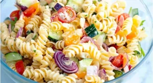 A clear glass bowl holds a fully mixed cold pasta salad recipe featuring rotini pasta, tomatoes, cucumbers, and a creamy dressing, ready to be served.