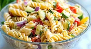 A close-up of a cold pasta salad recipe in a clear glass bowl, highlighting rotini pasta, cherry tomatoes, cucumbers, red onion, and a creamy dressing with fresh herbs.