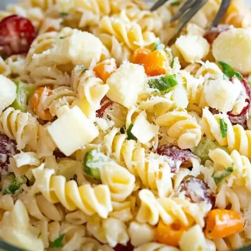 A clear glass bowl contains a partially mixed cold pasta salad recipe with rotini pasta, mozzarella, tomatoes, carrots, cucumbers, and fresh herbs. A silver fork is visible within the salad.