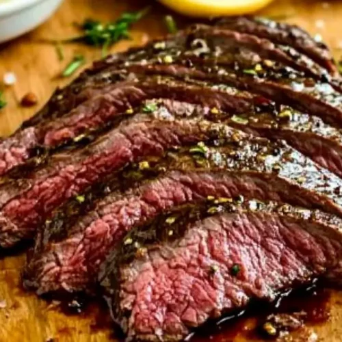 Slices of juicy flank steak with visible grill marks and pink center, seasoned with herbs and pepper, on a wooden cutting board with a side of sauce. Juicy Flank Steak Recipe.