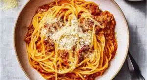 A creamy white bowl filled with steaming instant pot spaghetti topped with a rich meat sauce and grated Parmesan cheese, set on a light grey textured surface with a sprinkle of extra cheese and cutlery nearby.