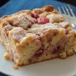 A slice of golden brown rhubarb coffee cake recipe dusted with powdered sugar sits on a white plate next to two silver forks, on a blue cloth napkin.