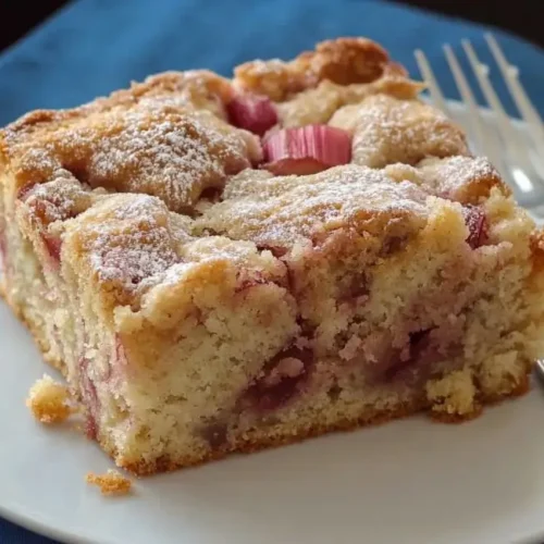 A slice of golden brown rhubarb coffee cake recipe dusted with powdered sugar sits on a white plate next to two silver forks, on a blue cloth napkin.