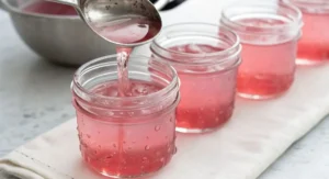 Clear liquid being poured from a spoon into small jars of pink rhubarb jelly.