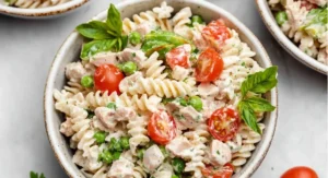 A close-up, overhead shot of a white bowl containing a tuna pasta salad recipe, including spiral pasta, tuna chunks, green peas, halved cherry tomatoes, and fresh basil leaves, all coated in a creamy dressing.