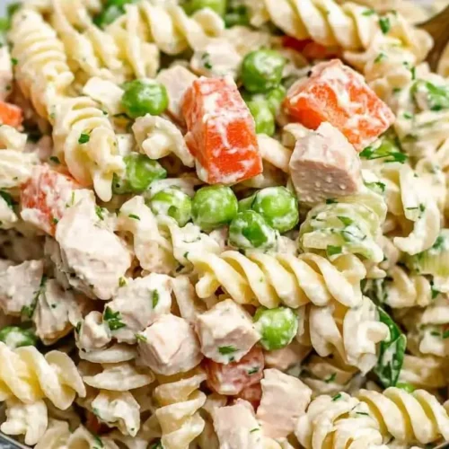 An angled, close-up view of a clear glass bowl filled with a tuna pasta salad recipe, showcasing spiral pasta, tuna chunks, diced carrots, green peas, and a creamy white dressing, mixed together.