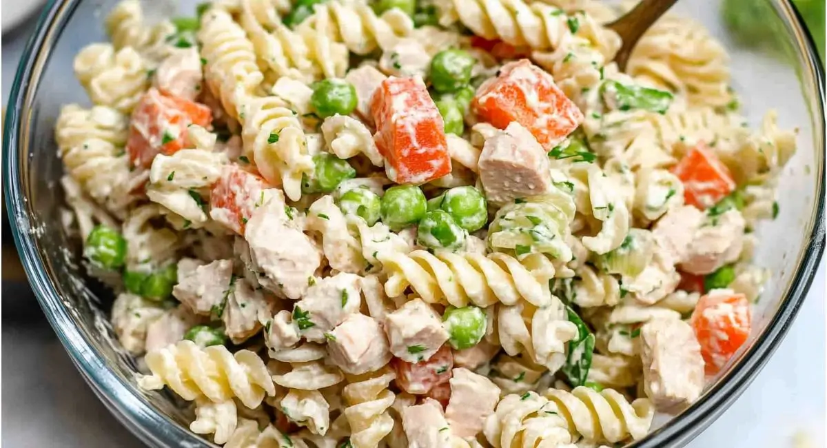 An angled, close-up view of a clear glass bowl filled with a tuna pasta salad recipe, showcasing spiral pasta, tuna chunks, diced carrots, green peas, and a creamy white dressing, mixed together.