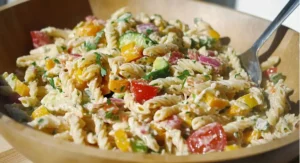 A wooden bowl filled with vegan pasta salad recipe containing rotini pasta, colorful vegetables, and a creamy dressing, with a silver fork resting in the salad.