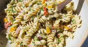 A white bowl containing a vegan pasta salad recipe with rotini pasta, colorful bell peppers, red onion, parsley, and a creamy dressing, being stirred with a wooden spoon.